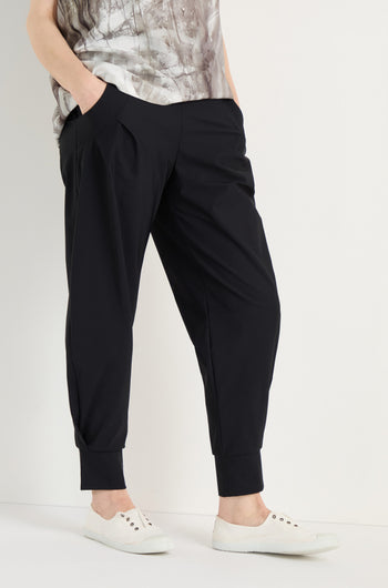 Person stands with hands in pockets, wearing Tapered Cuff Trousers made from stretch fabric, a patterned top, and white slip-on sneakers against a plain background.