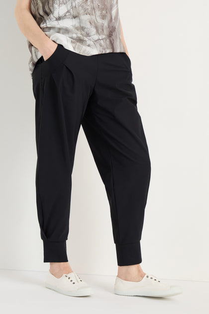 Person stands with hands in pockets, wearing Tapered Cuff Trousers made from stretch fabric, a patterned top, and white slip-on sneakers against a plain background.