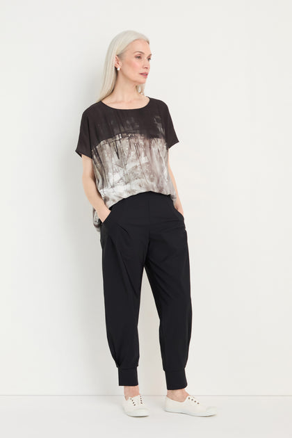 A woman with long gray hair stands against a white wall, hands in her pockets, wearing a loose printed top, white sneakers, and Tapered Cuff Trousers.