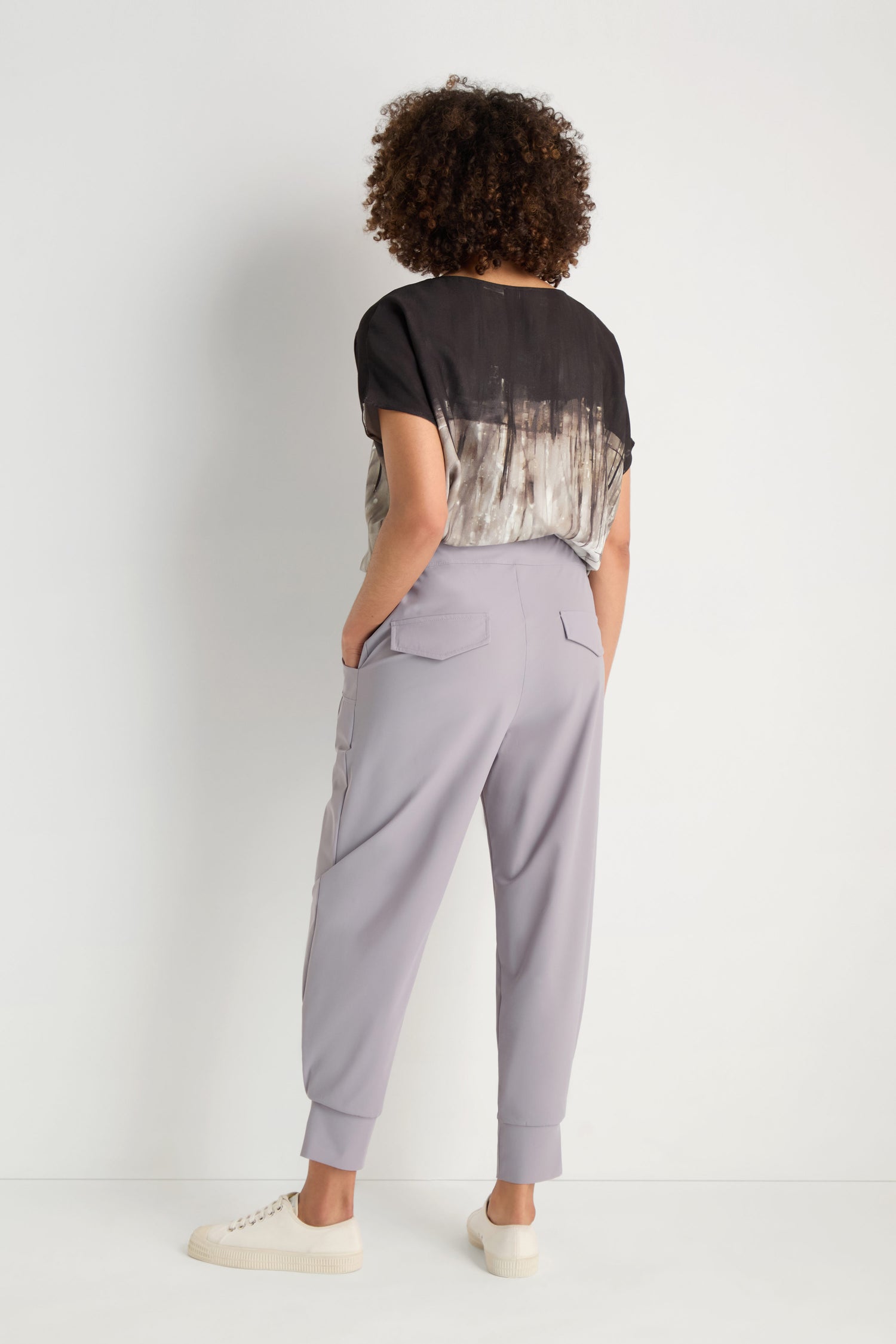 A person stands facing away, wearing a loose black-and-white patterned top, Tapered Cuff Trousers in light gray, and white sneakers against a plain white background.