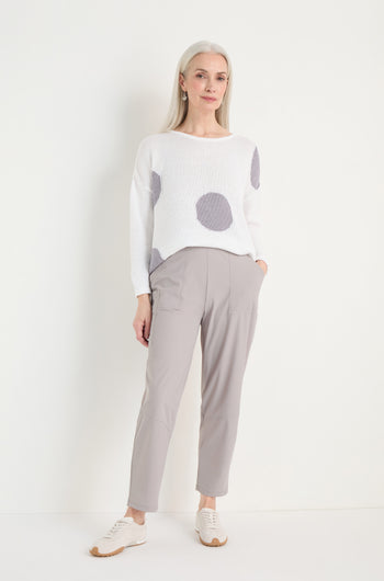 A woman stands against a white wall wearing a white sweater with large gray polka dots, light gray Tapered Trousers in stretch fabric, and white sneakers.