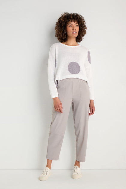 A woman stands against a plain white wall wearing a white sweater with large gray polka dots, Tapered Trousers in light gray cropped length, and white sneakers.