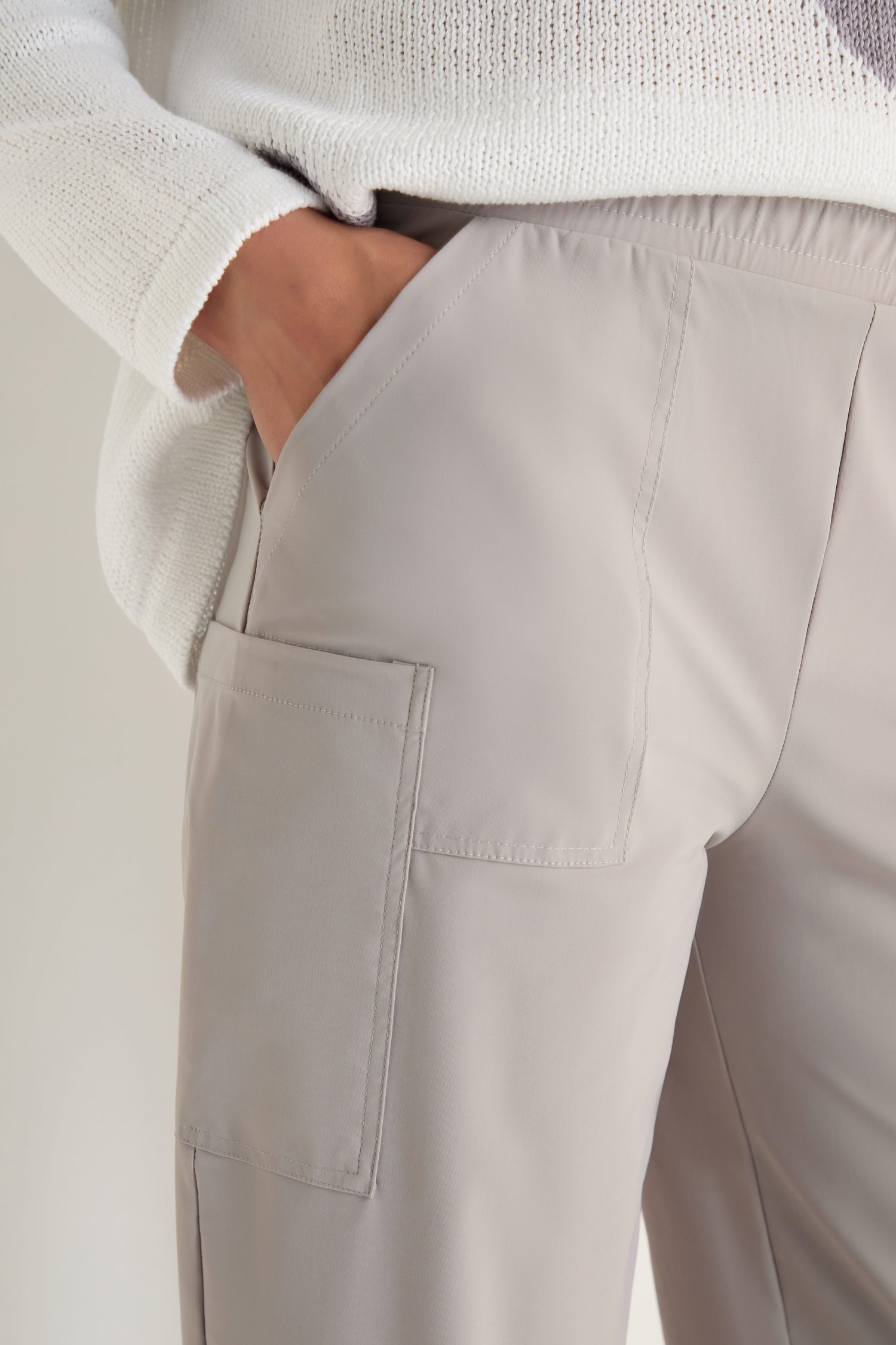Person wearing Tapered Trousers in light beige with a side pocket and stretch fabric for comfort, styled with a white textured sweater. The trousers have a modern, cropped cut and the person has one hand in the pocket.