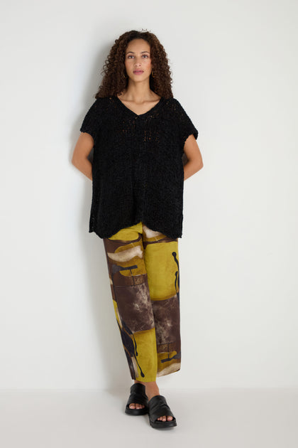 A woman stands against a white wall wearing a relaxed silhouette V-Neck Cotton Blend Black Knit, wide-leg abstract print pants in earth tones, and black slide sandals.