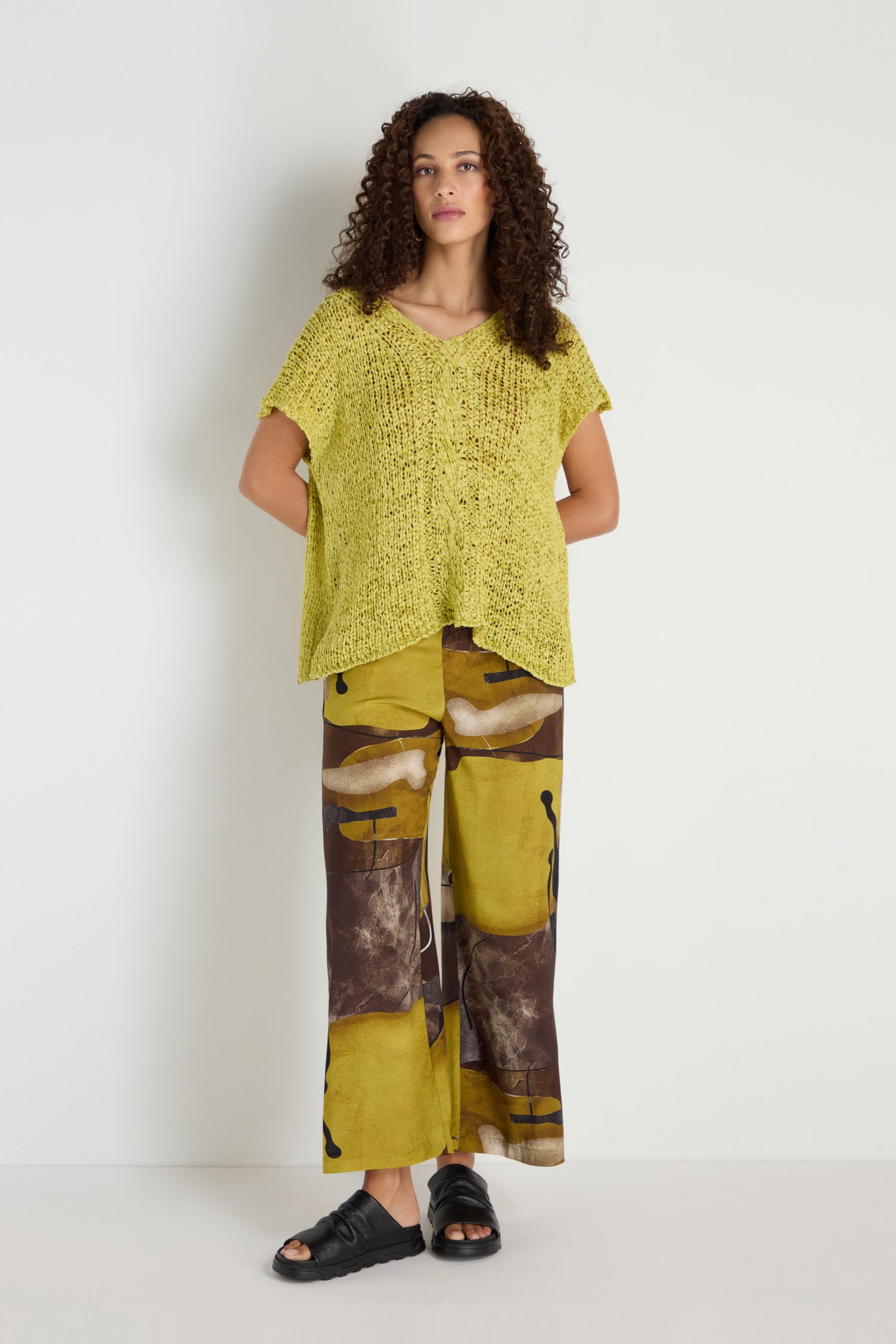A woman stands against a white wall wearing a loose V-Neck Cotton Blend Knit in yellow, wide-leg abstract yellow and brown pants, and black sandals.