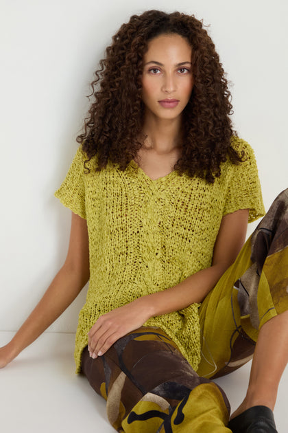 A woman with curly hair wears the V-Neck Cotton Blend Knit in yellow and patterned brown-and-yellow pants, seated against a white background.