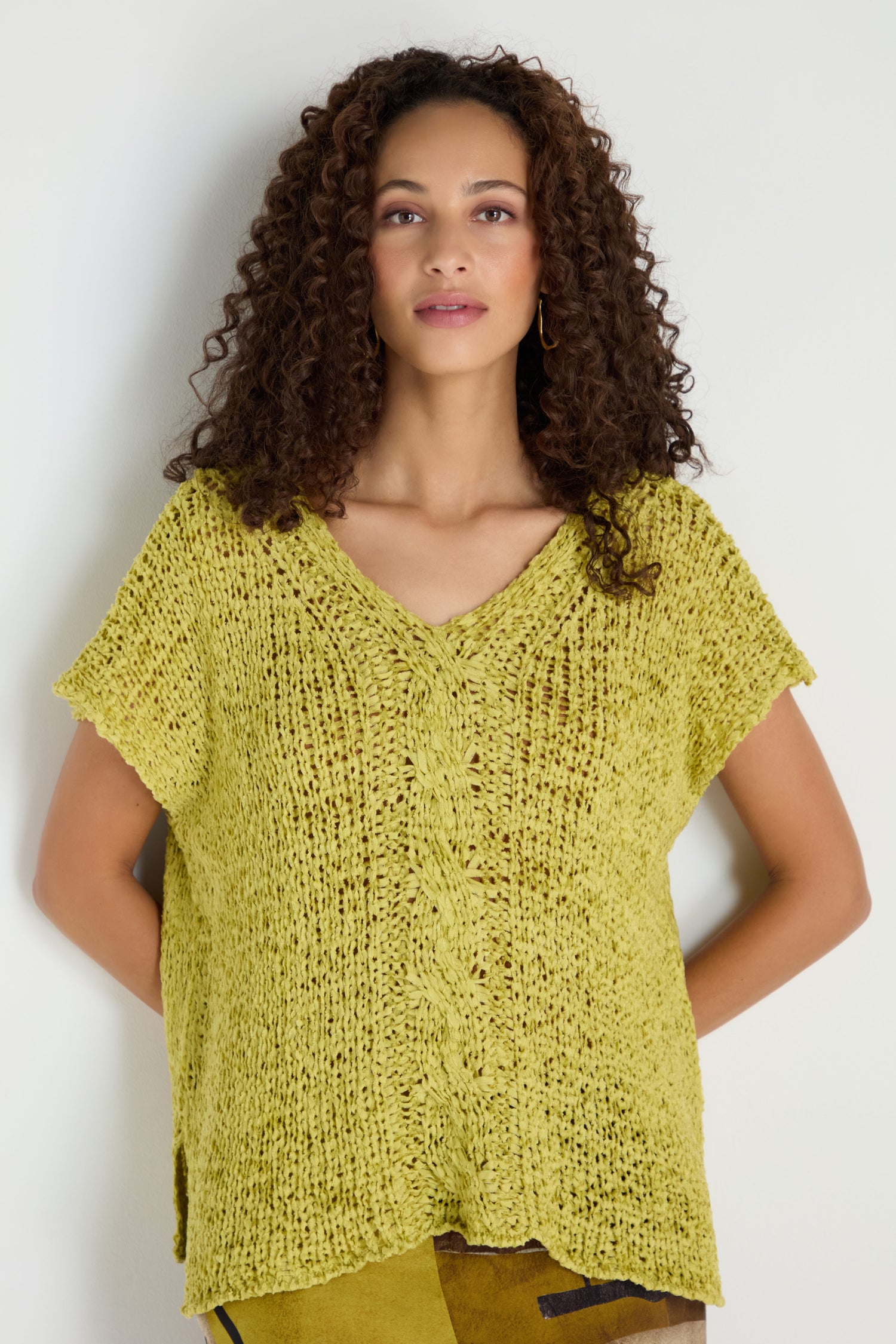 A woman with curly hair wears the V-Neck Cotton Blend Knit, a loose-fitting, short-sleeve yellow top with front cable detail, standing against a plain light background.