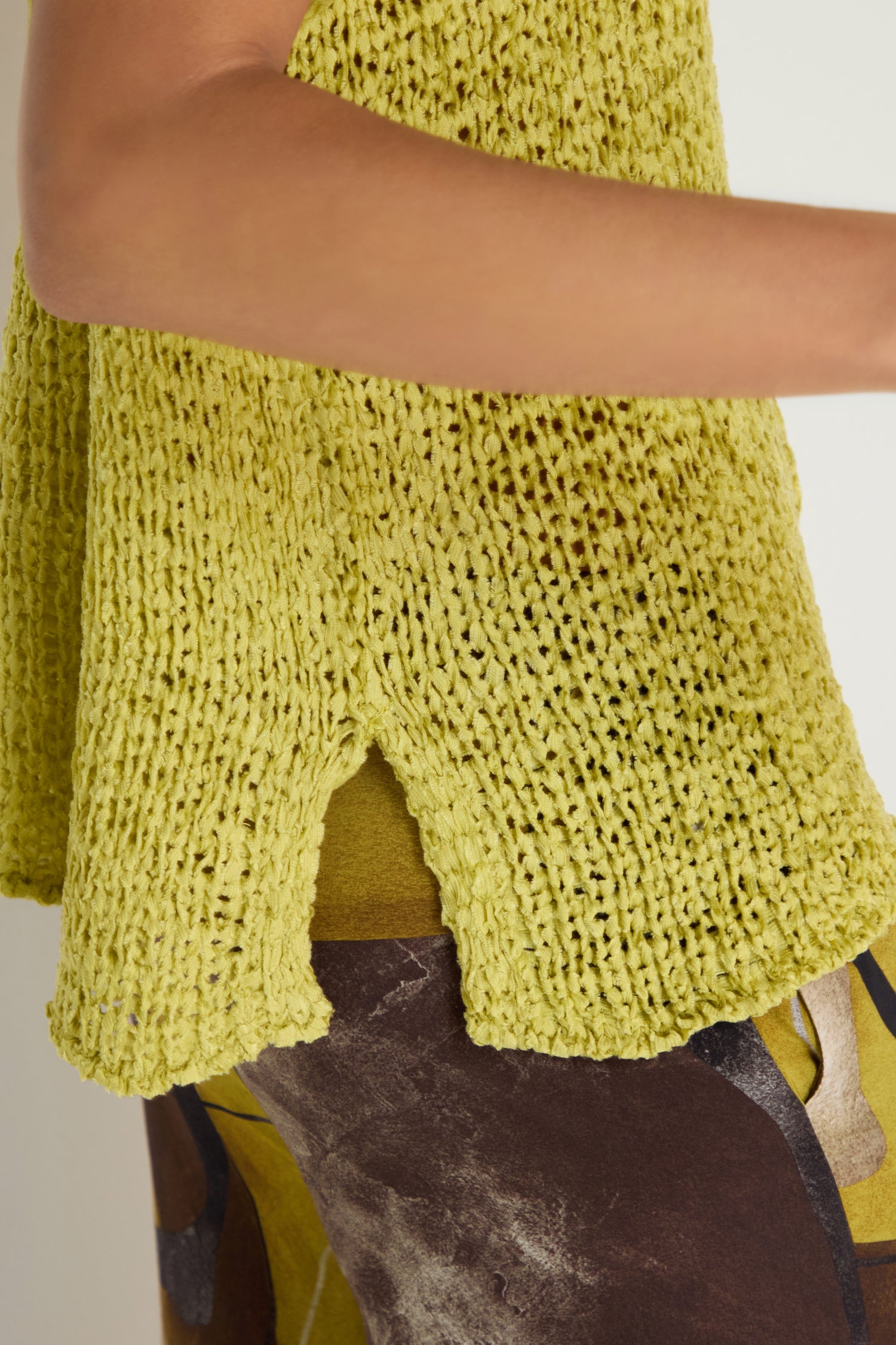 A close-up of a person wearing the V-Neck Cotton Blend Knit in yellow-green, featuring a loose-knit sleeveless design and small side slit, paired with abstract brown and yellow patterned pants.