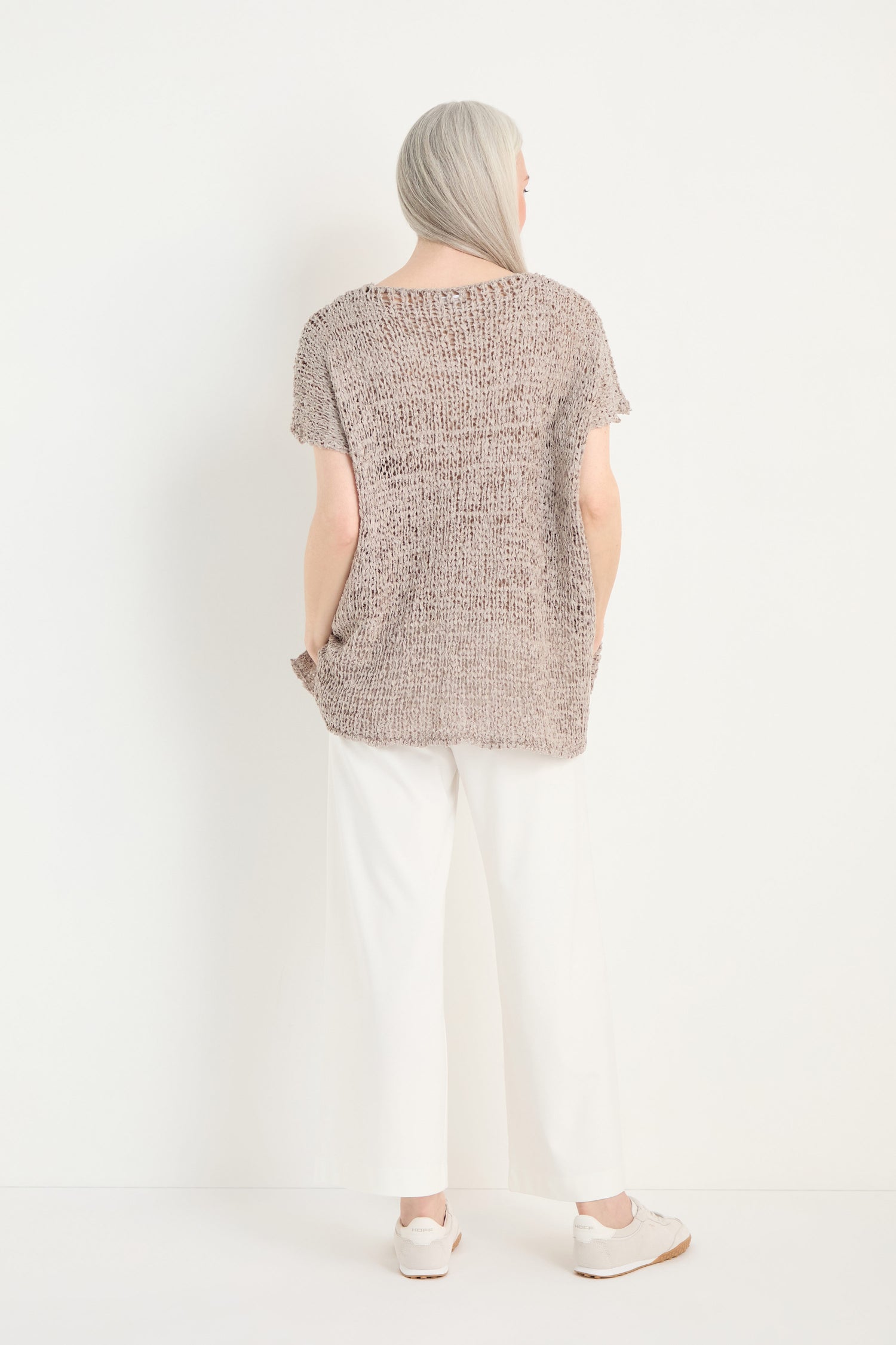 An older woman with long gray hair faces a white wall, wearing the V-Neck Cotton Blend Knit in beige, paired with white pants and light-colored shoes.