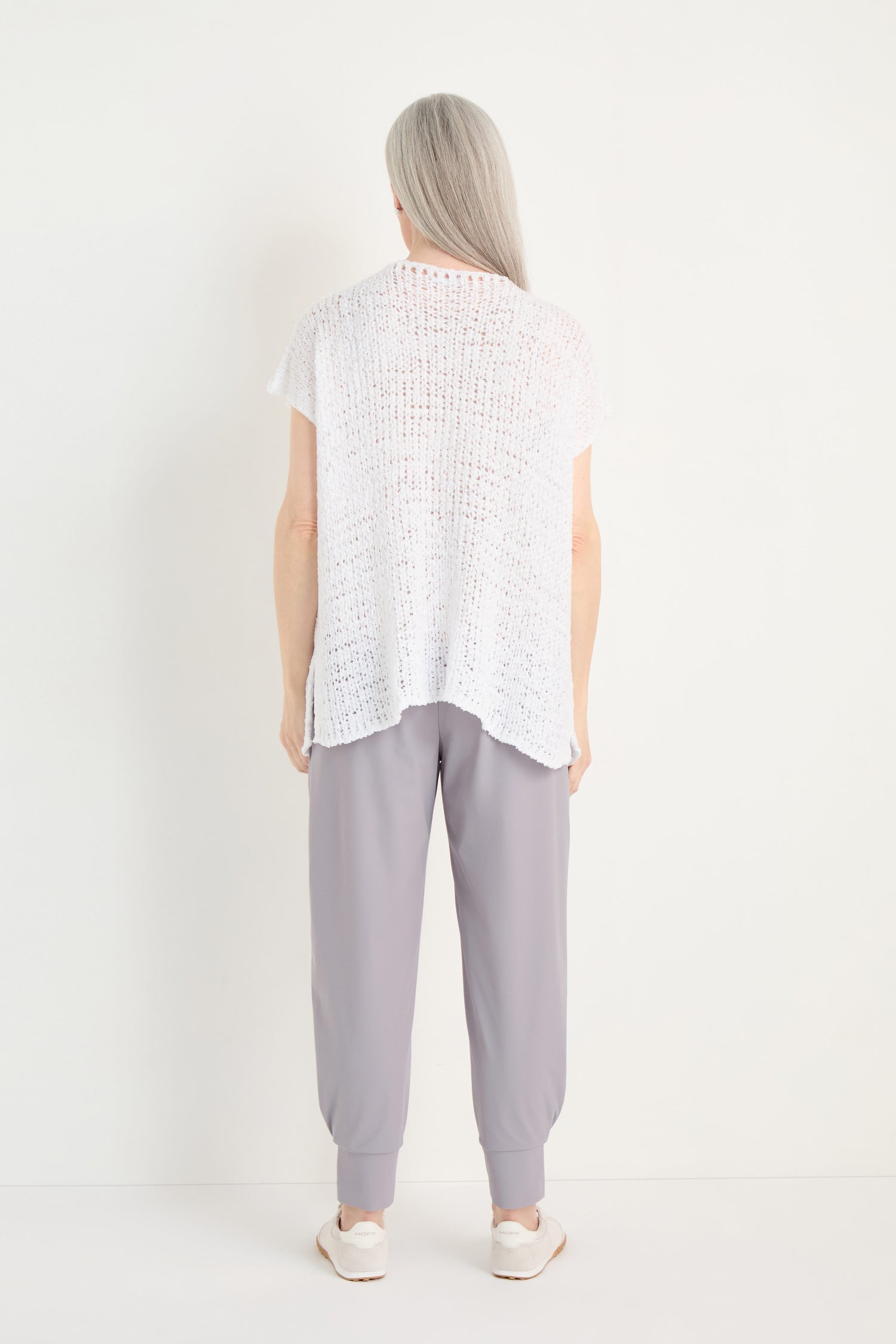A person with long gray hair, seen from behind, is wearing a loose white V-Neck Cotton Blend Knit top, light gray pants, and white shoes, standing against a plain white background.