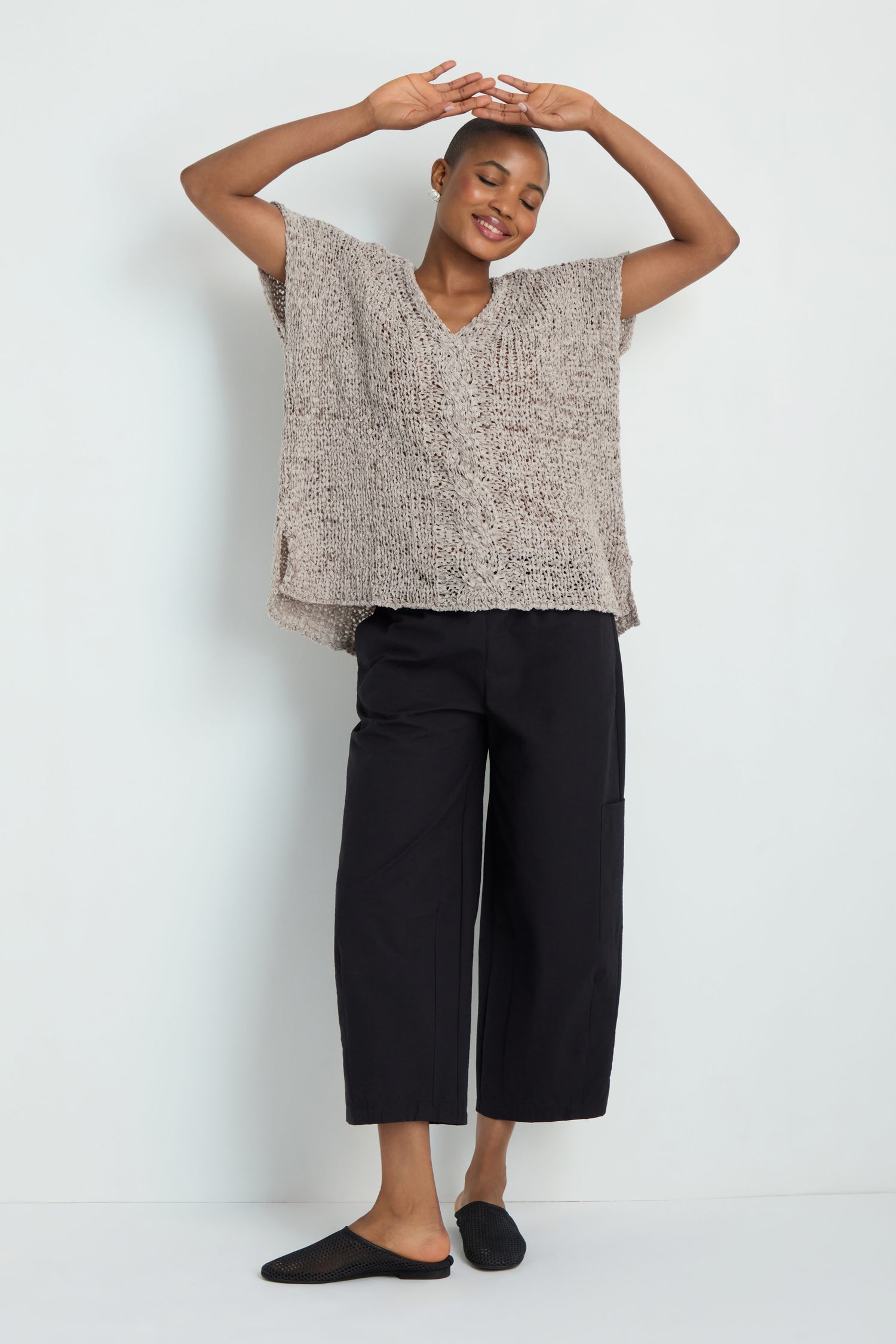Person stands against a plain background, smiling with arms raised and eyes closed, wearing the V-Neck Cotton Blend Knit in beige, black wide-leg pants, and black slip-on shoes.