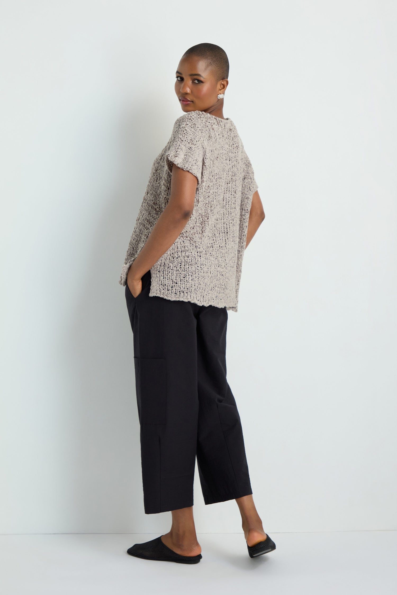 A person stands against a white background wearing the V-Neck Cotton Blend Knit in beige, paired with black cropped pants and black slip-on shoes, hands in pockets and body turned slightly to the side.