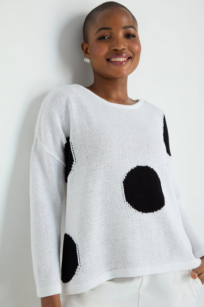 A woman with long gray hair wears the Spot Cotton Knit, a white sweater with a bold spot pattern, paired with black pants, standing against a plain white background.