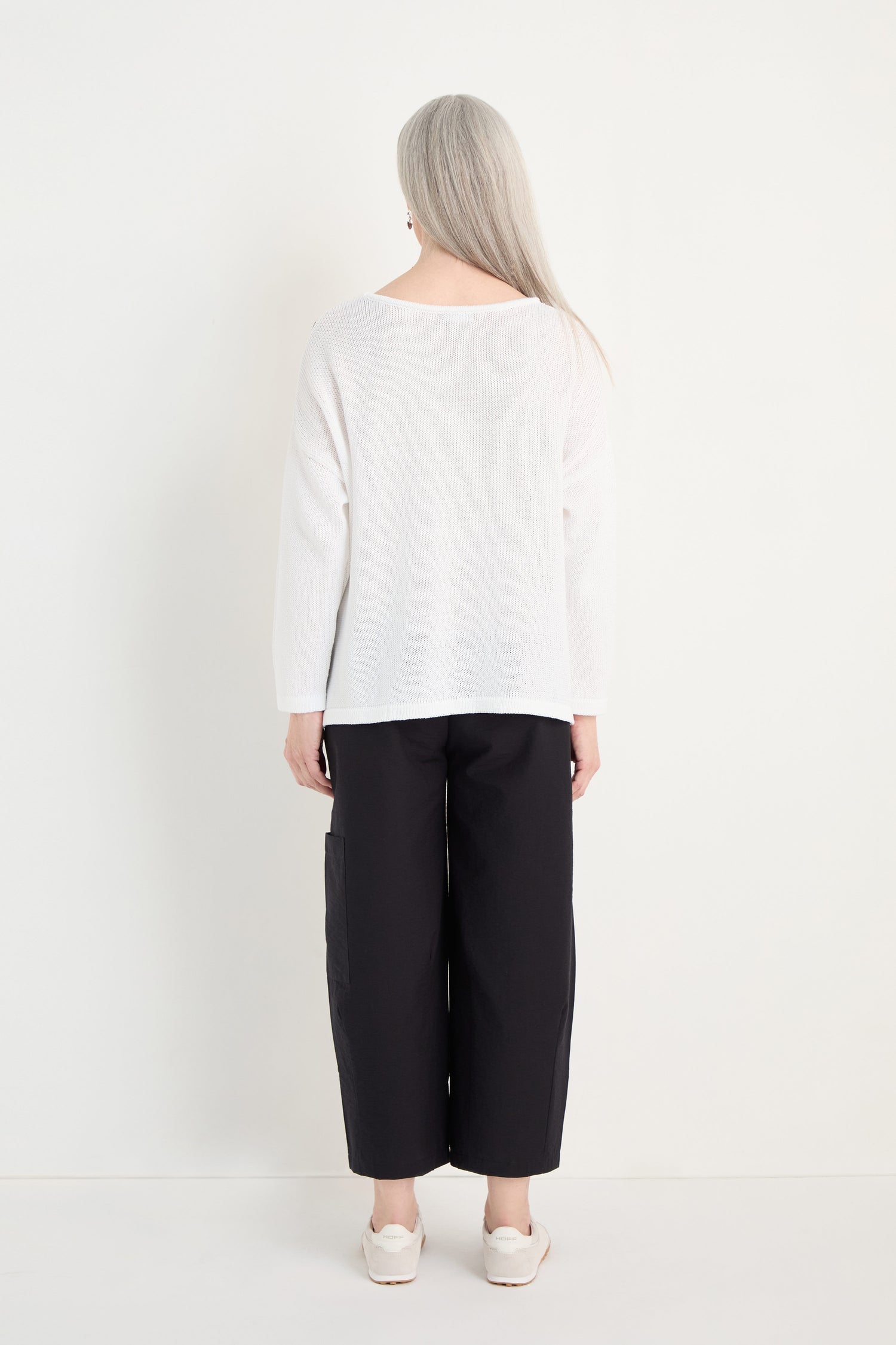 A person with long gray hair faces away, wearing the Spot Cotton Knit sweater in white, paired with black wide-leg pants and white shoes, all set against a plain white background.
