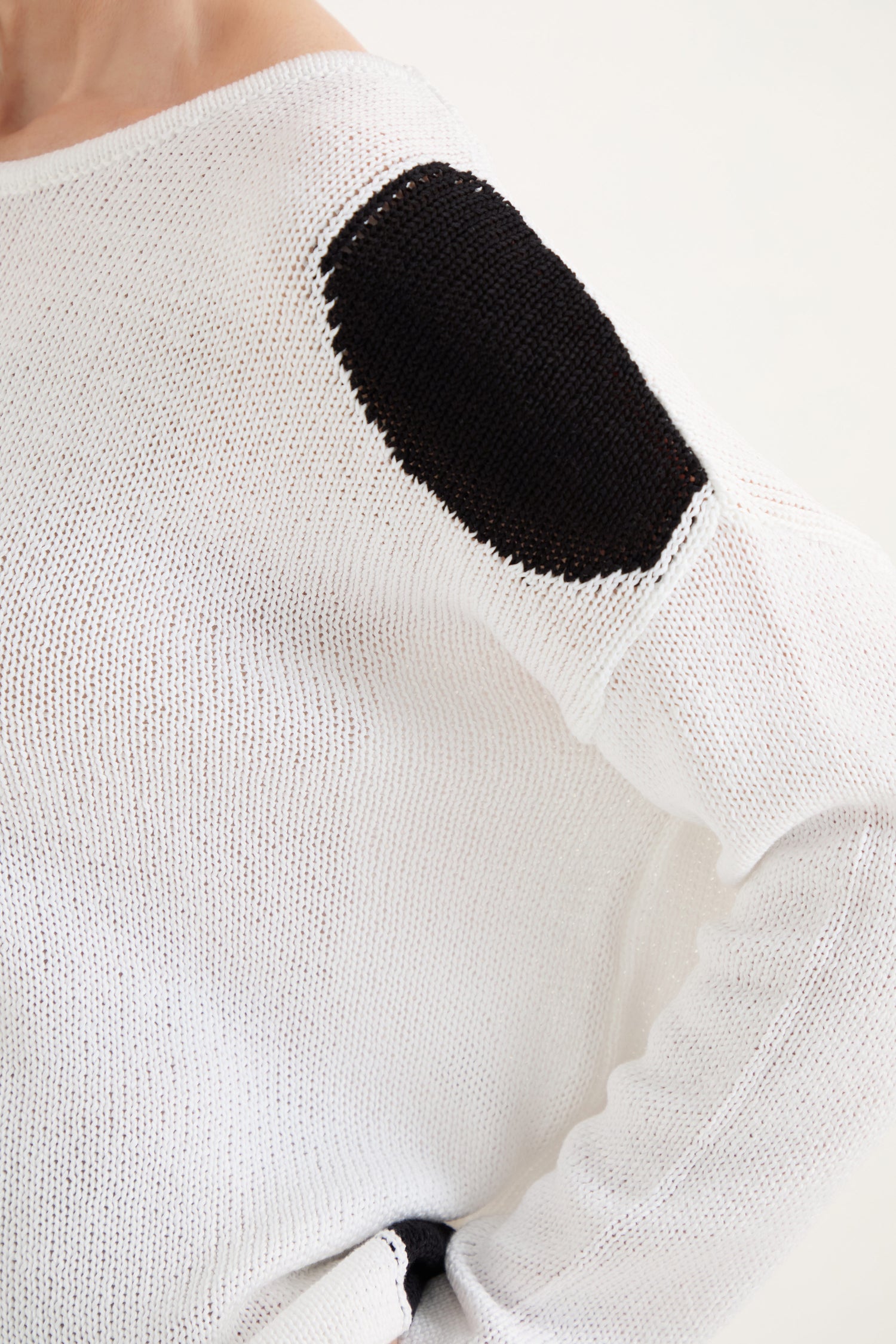 Close-up of the Spot Cotton Knit sweater in white organic cotton with a black oval shoulder patch; features a relaxed fit and one arm bent at the elbow.