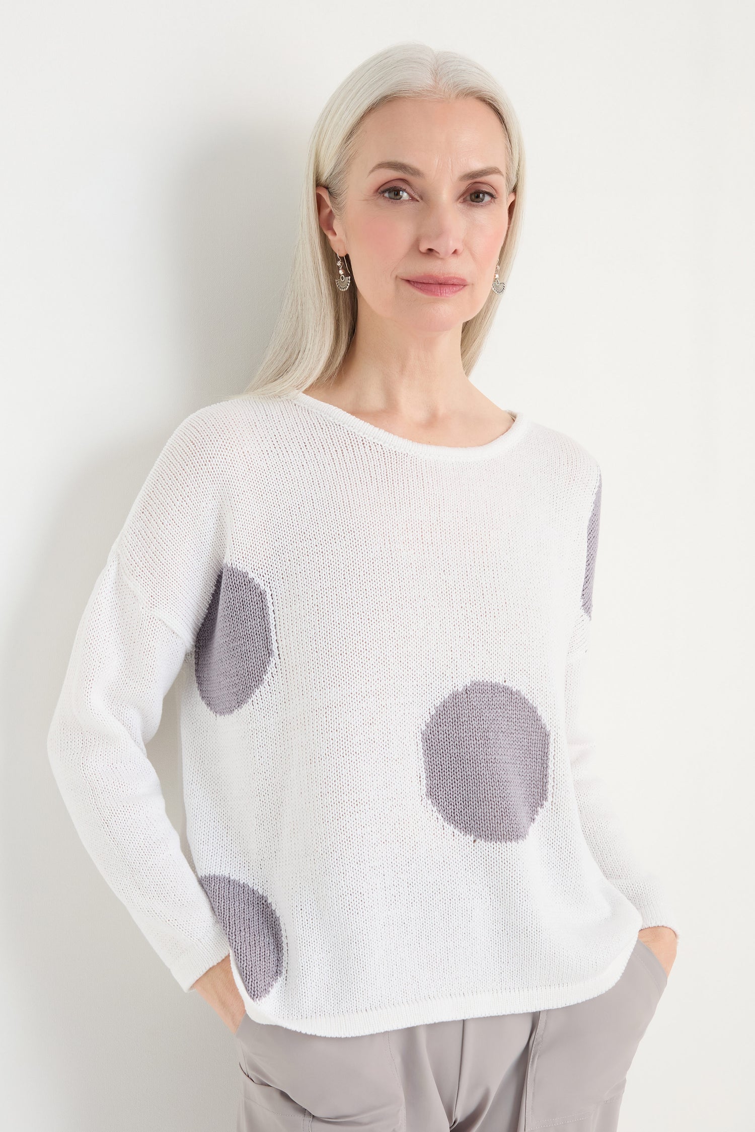 A woman with long gray hair wears the Spot Cotton Knit, a relaxed white sweater with bold spots, paired with light gray pants, standing against a white background.