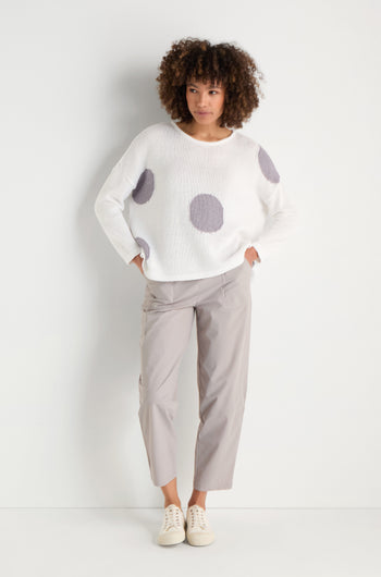 A woman stands against a white background, wearing the Spot Cotton Knit sweater in white with a bold spot pattern, paired with light gray cropped pants and white sneakers.