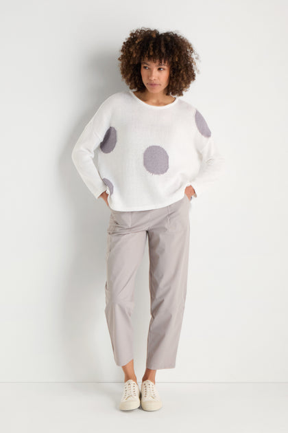 A woman stands against a white background, wearing the Spot Cotton Knit sweater in white with a bold spot pattern, paired with light gray cropped pants and white sneakers.