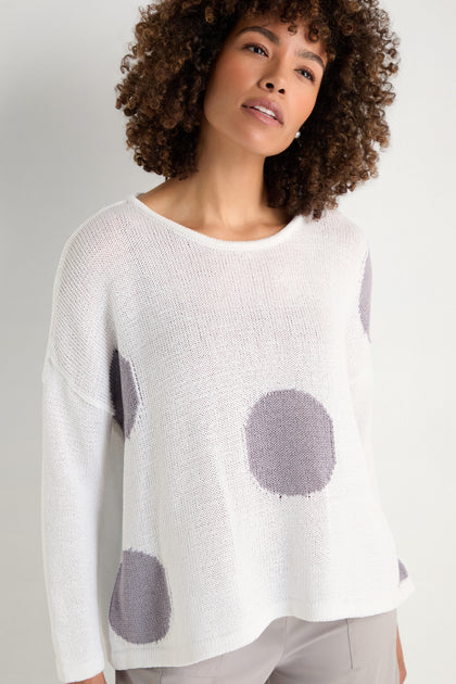 A woman stands against a plain background wearing the Spot Cotton Knit—a relaxed silhouette white organic cotton sweater featuring large gray polka dots.