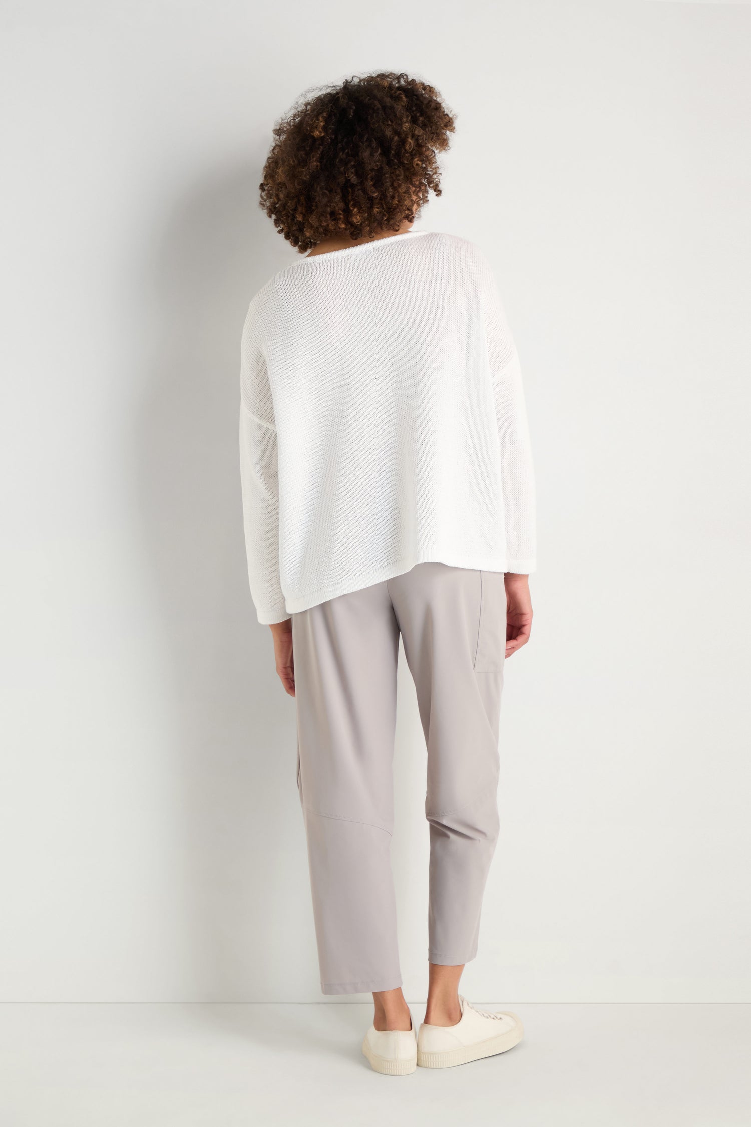 A person stands facing a white wall, wearing the Spot Cotton Knit sweater in organic cotton, paired with light gray pants and white shoes.