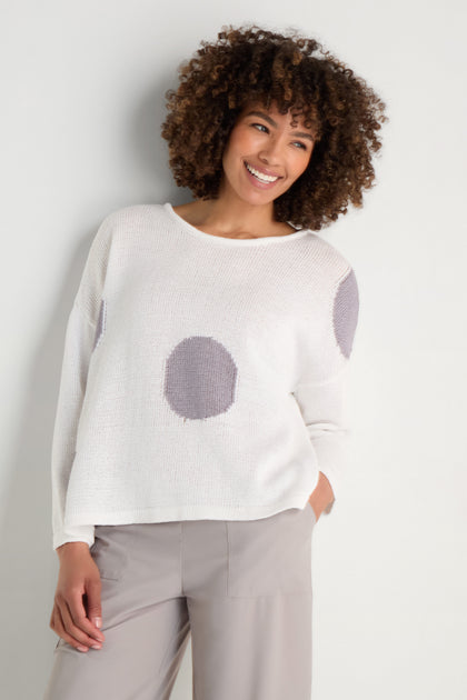 A woman smiles in a relaxed silhouette, wearing the Spot Cotton Knit—an organic cotton sweater with a bold spot pattern—paired with light gray pants against a plain background.
