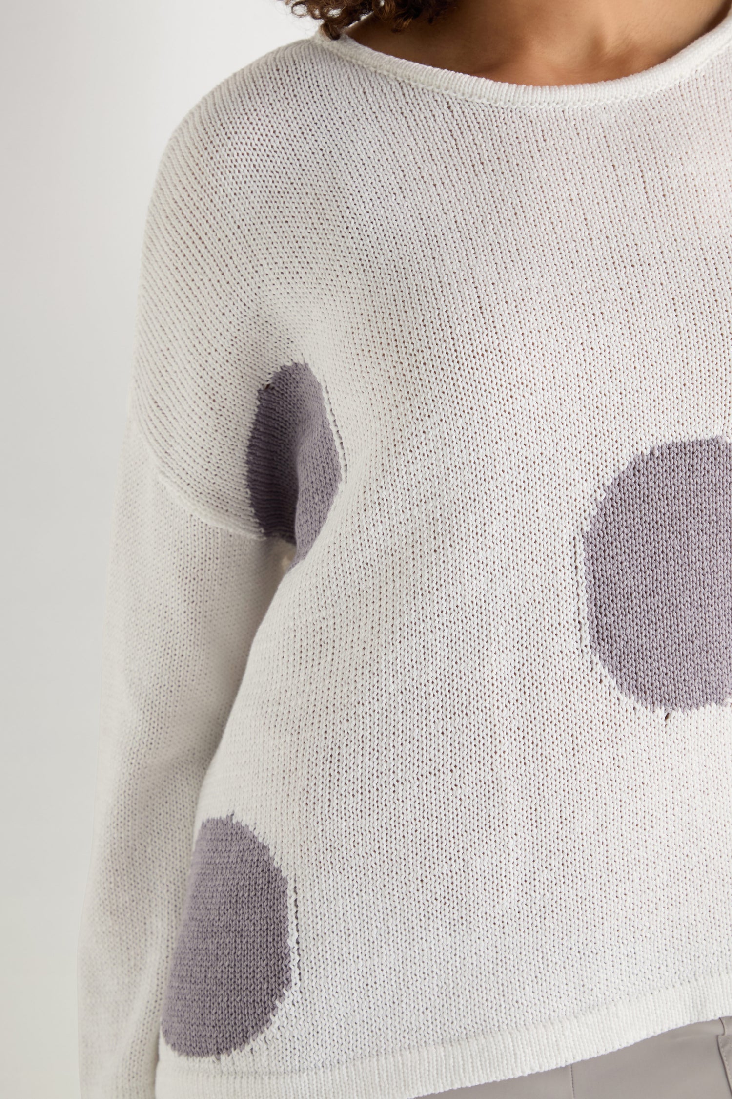 A person wears the Spot Cotton Knit, an organic cotton white sweater with a bold spot pattern; upper body partially shown against a plain background.