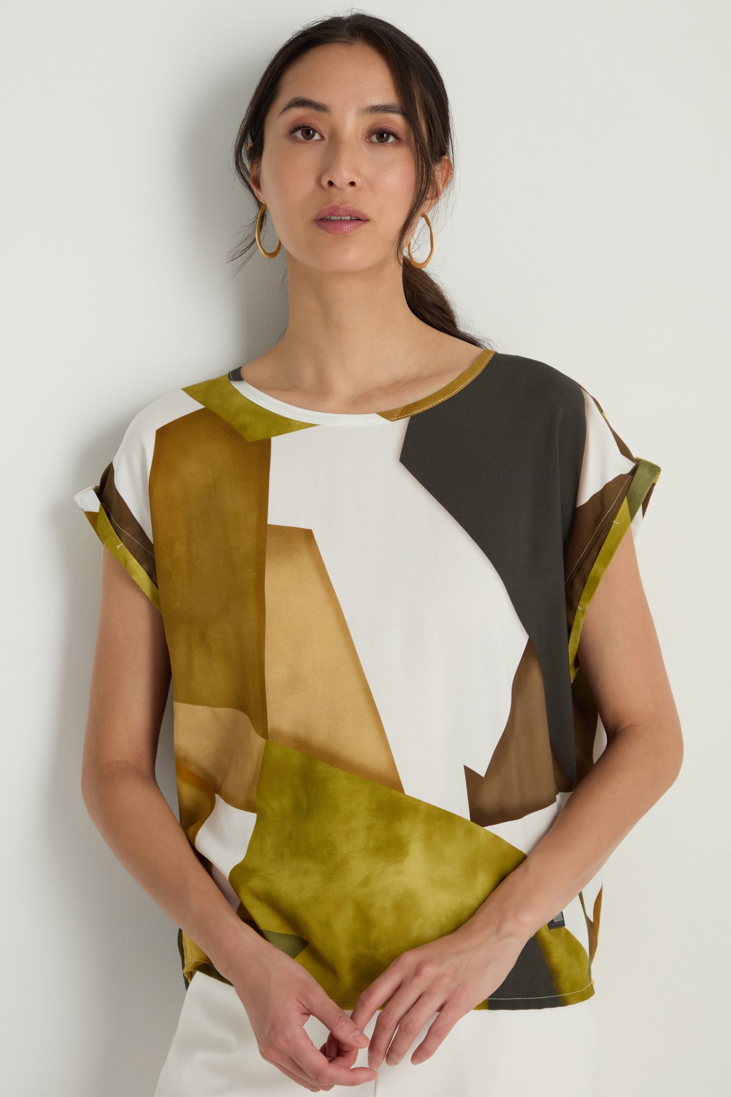 A woman stands against a white wall wearing the Collage Print Top, featuring a relaxed fit, soft viscose fabric, an abstract green, brown, and white geometric design, paired with gold hoop earrings.