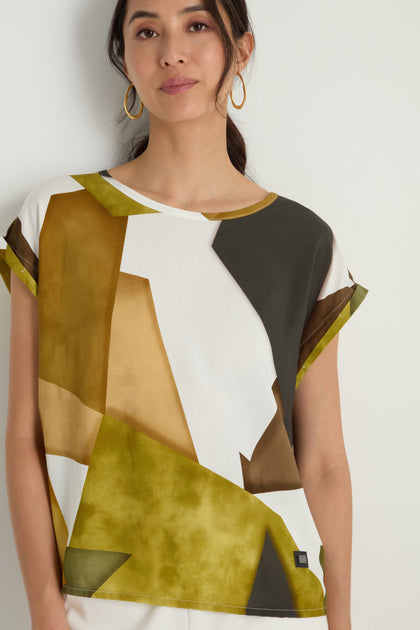 A woman stands against a white background wearing the Collage Print Top—a relaxed fit, short-sleeved top featuring olive, brown, white, and black collage prints and crafted from soft viscose.