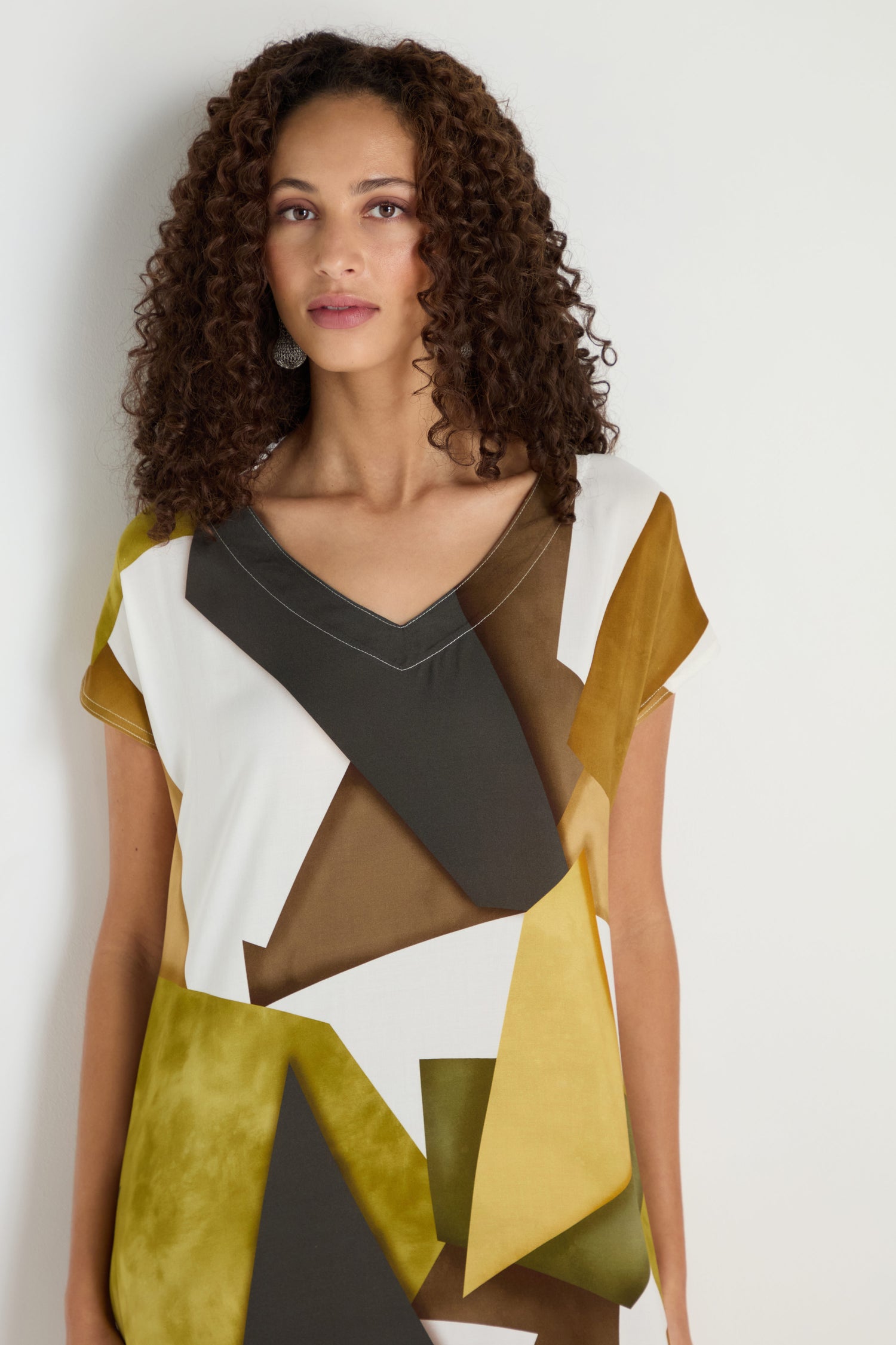A woman with curly hair stands against a plain white background, wearing a short-sleeved Collage Print V-Neck Dress in yellow, brown, black, and white.