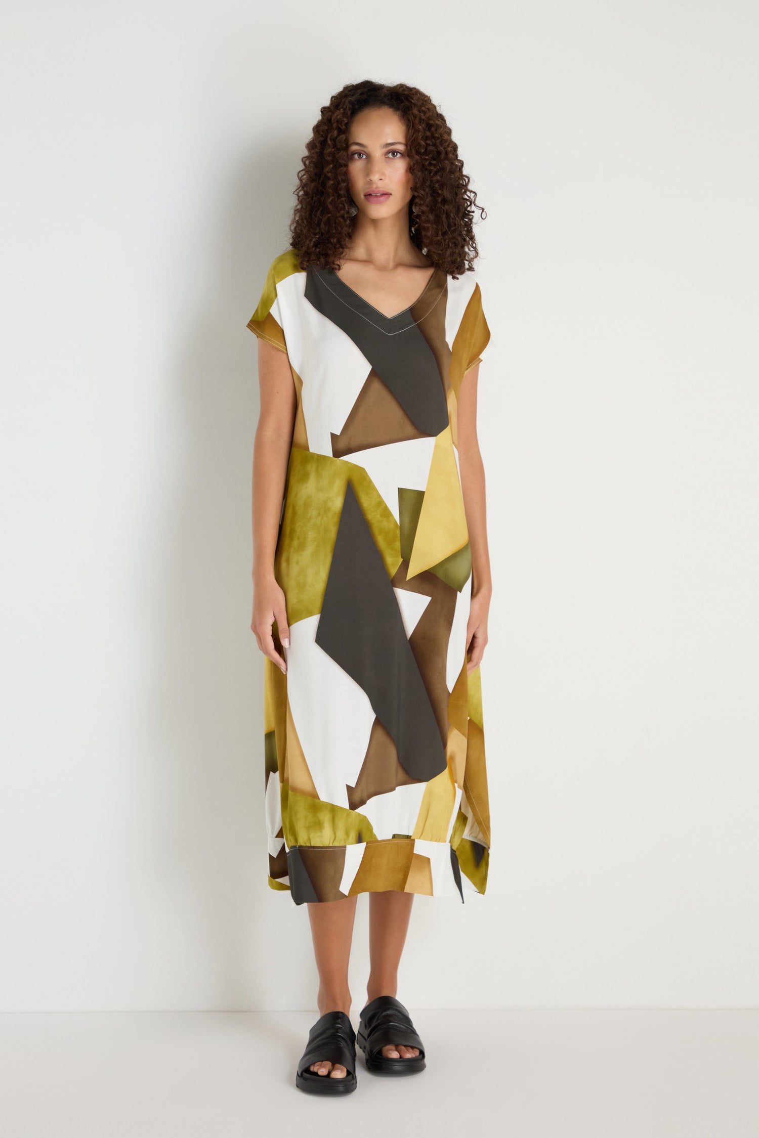 A woman stands against a plain white wall wearing a loose, knee-length Collage Print V-Neck Dress featuring an abstract geometric pattern in olive, black, white, and brown, paired with black sandals.