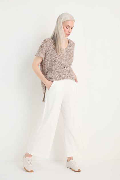 A woman with long gray hair stands against a plain white background, hands in pockets, wearing Wide Leg Pocket Trousers with an elastic waist, a loose taupe sweater, and white sneakers.