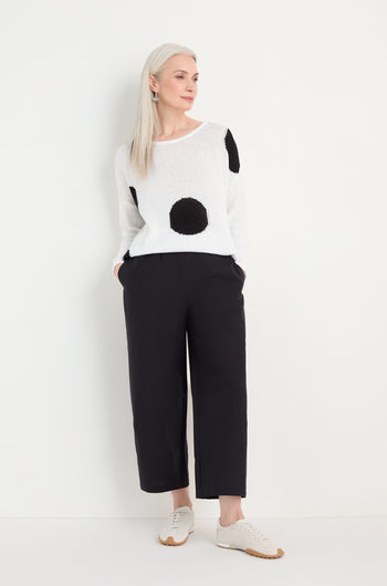 A woman stands against a white wall wearing a white sweater with large black dots, Wide Leg Cotton Trousers, and white sneakers.