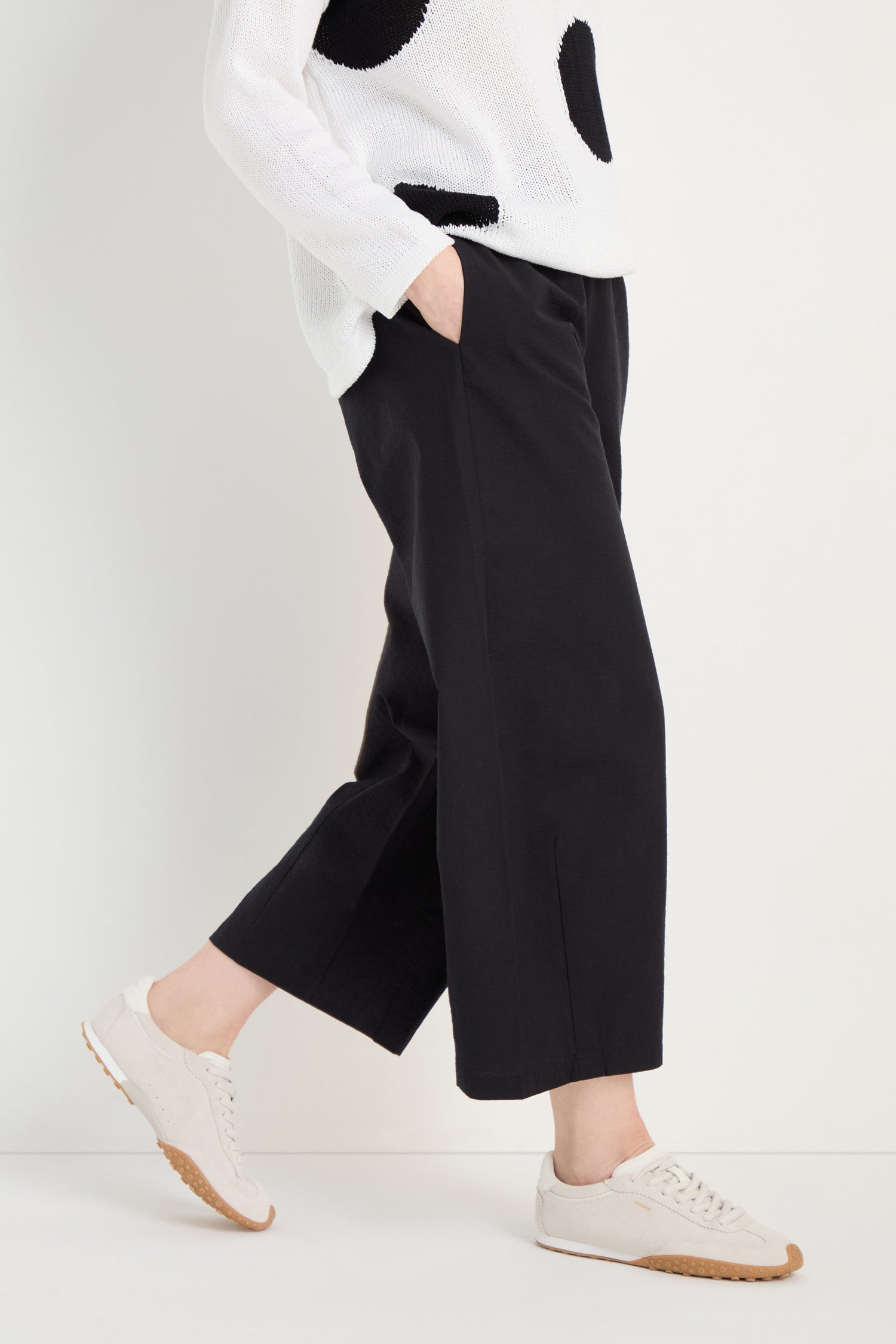 Person wearing Wide Leg Cotton Trousers in black, a white sweater with black spots, and beige sneakers, standing with one hand in their pocket against a plain background.