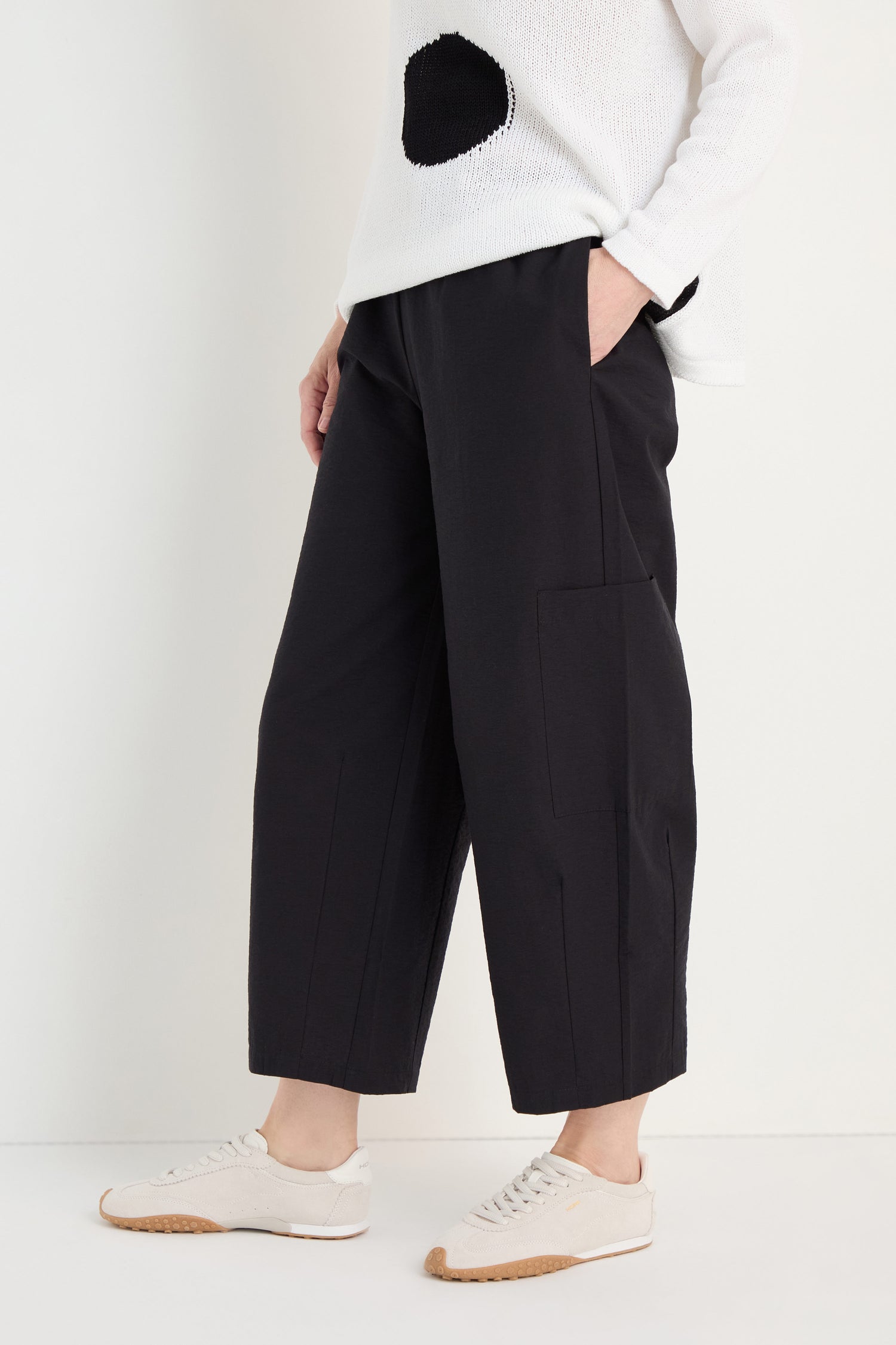 A person wearing a white sweater with a black circle, Wide Leg Cotton Trousers, and white sneakers stands with one hand in their pocket against a light background.