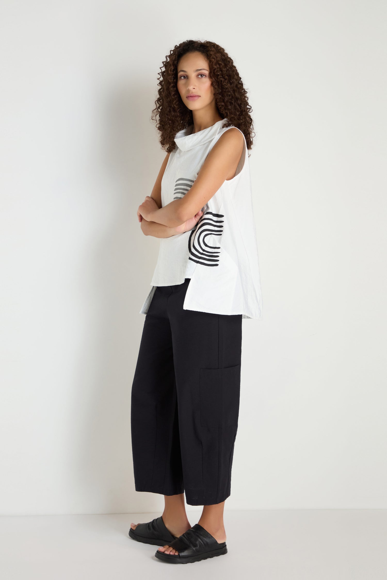 A woman with curly hair stands against a white background, wearing the Roll Collar Step Hem Top with black geometric patterns, paired with black wide-leg pants and black sandals.