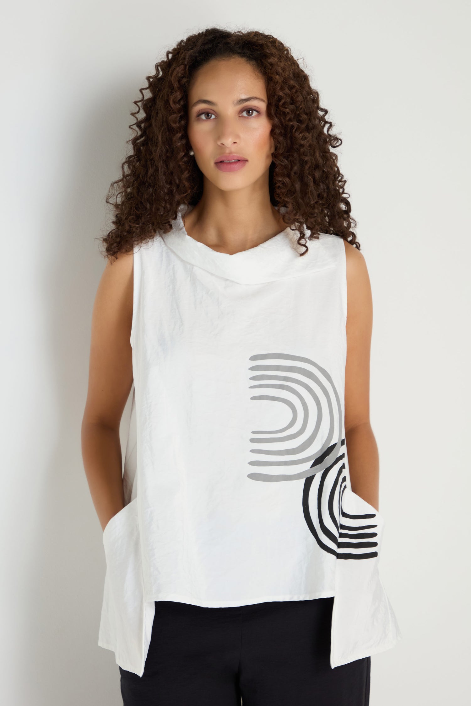 A woman with curly hair wears the Roll Collar Step Hem Top, a sleeveless white piece with black and gray geometric patterns, paired with black pants and standing against a plain light background.