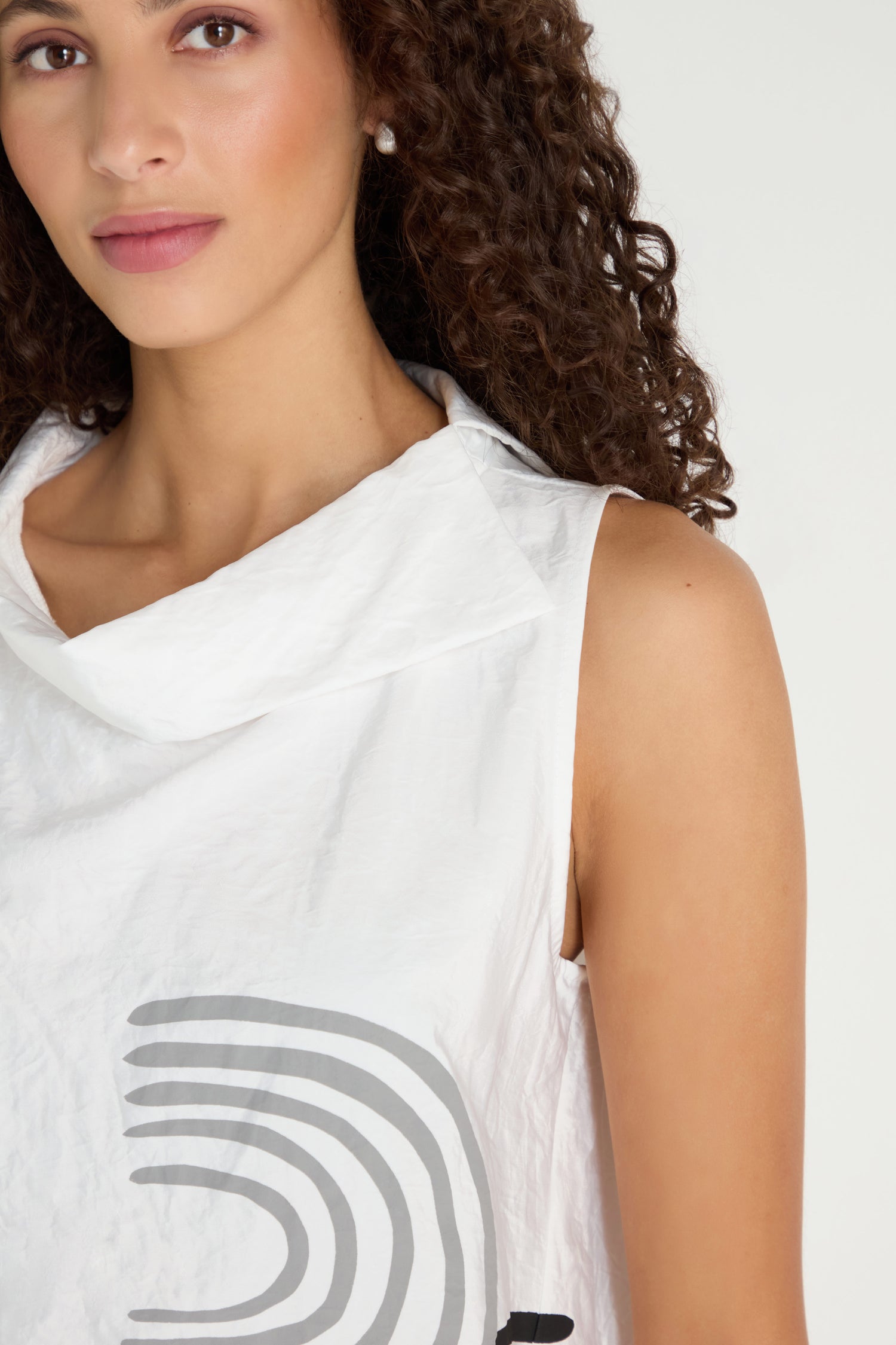 A woman with curly hair is shown from the shoulders up wearing the Roll Collar Step Hem Top, a sleeveless white top with a gray abstract graphic print.
