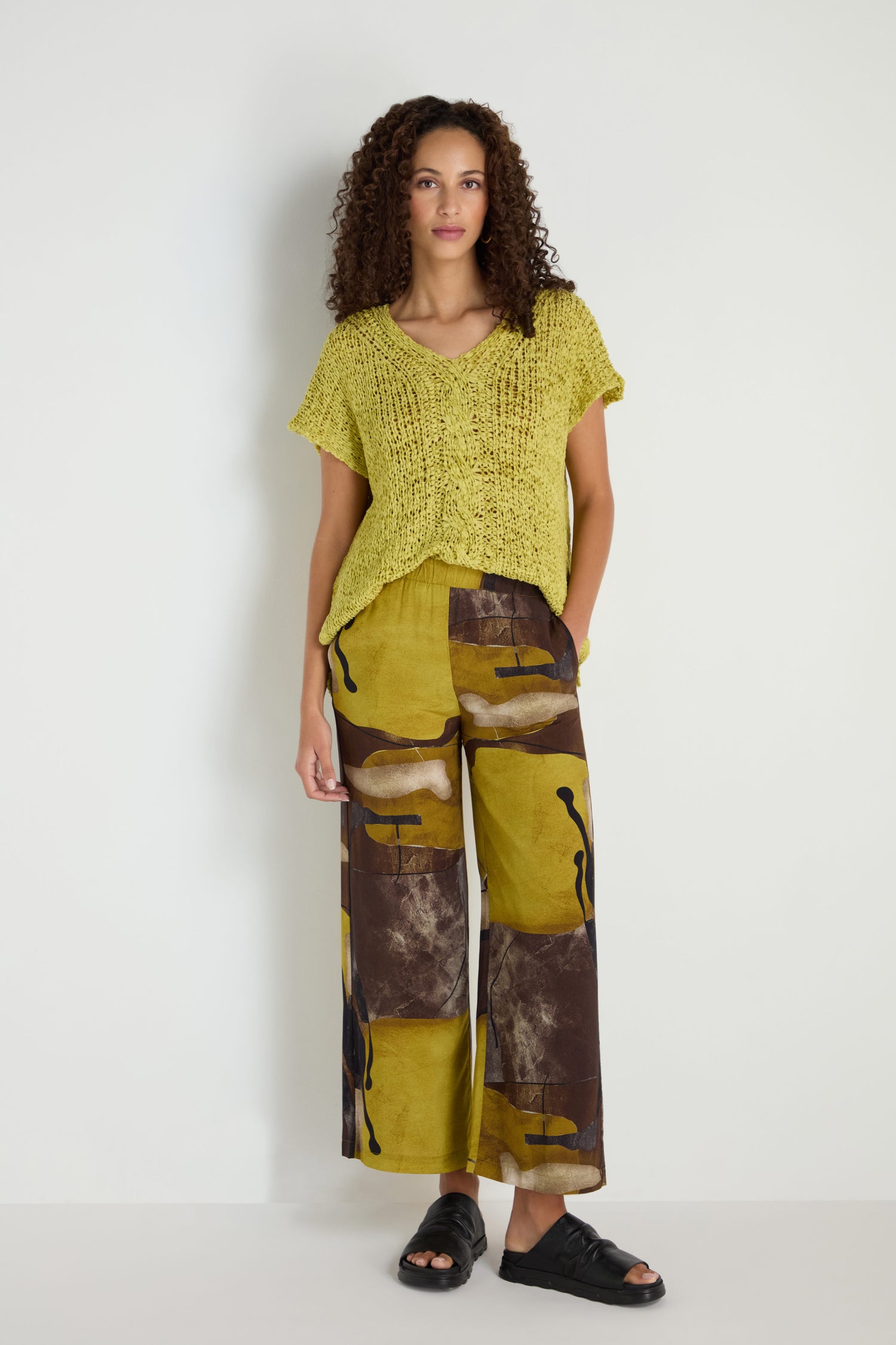 A woman stands against a white wall wearing Abstract Print Trousers in yellow and brown with a relaxed wide fit, paired with a loose yellow knit top and black sandals.