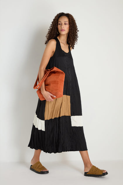 A woman stands against a plain white background wearing the Colour Block Pleated Dress in black, brown, and white, styled with a large rust tote bag and brown slide sandals.