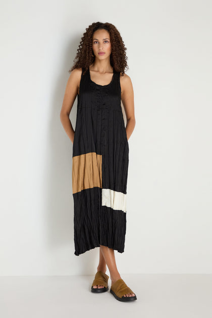 A woman stands against a white wall wearing the Colour Block Pleated Dress, a sleeveless black dress featuring tan and white accents, styled with brown sandals.