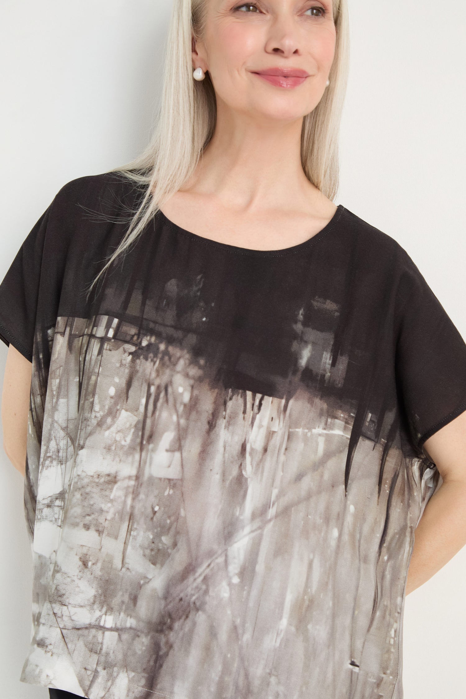 A woman with long gray hair wears the Abstract Print Easy Top in black and gray, smiling slightly while standing against a plain white background.