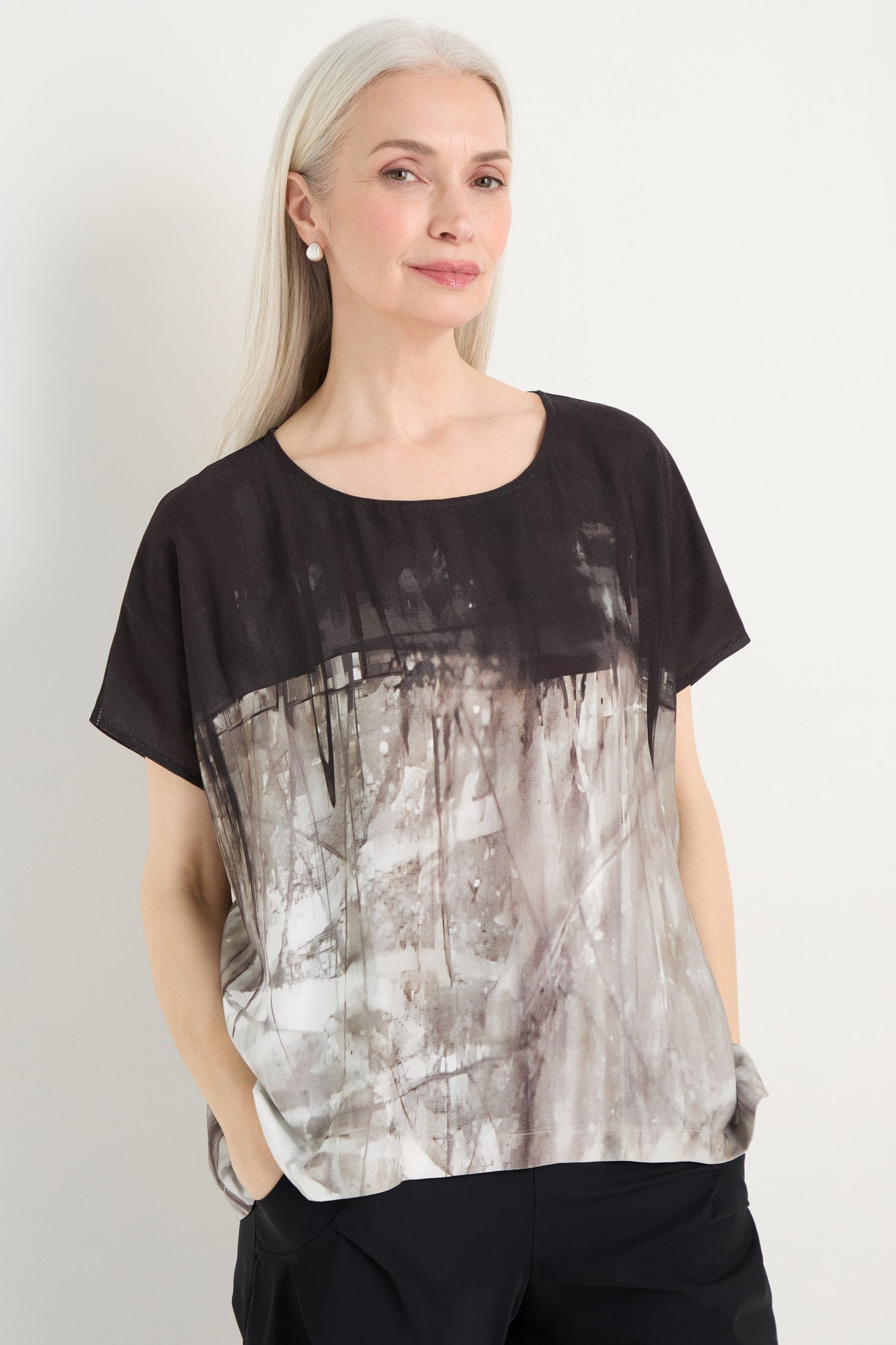 A woman with long gray hair wears the Abstract Print Easy Top in black and gray, paired with black pants, standing with her hands in her pockets against a plain background.