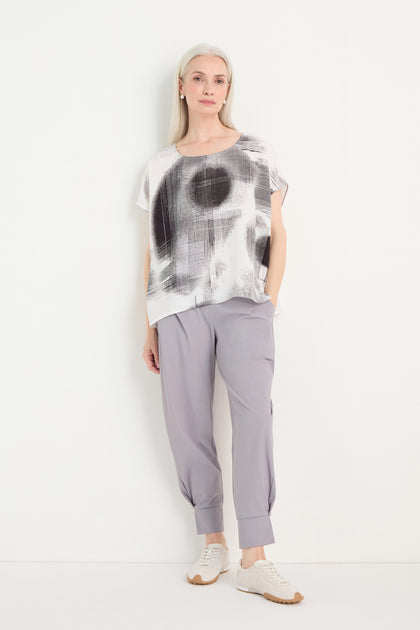 A woman stands against a white wall wearing a Shaded Print Top with a relaxed silhouette, light grey tapered pants, and white sneakers.