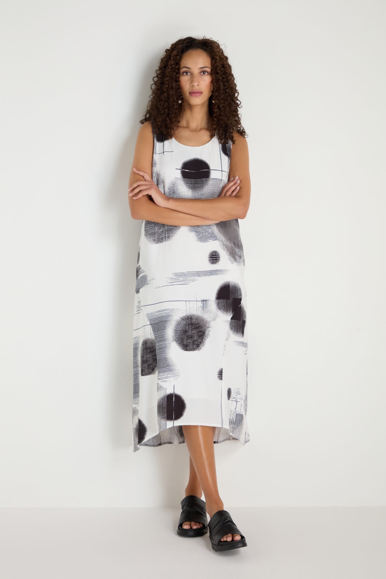 A woman stands against a white wall wearing the Shaded Print Flared Dress with black sandals, arms crossed and a neutral expression.