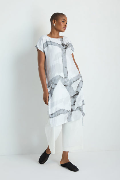 A woman with long white hair models the Brushstroke Print Long Tunic, featuring short sleeves and a knee-length hem, styled with black leggings and sandals against a plain white background.