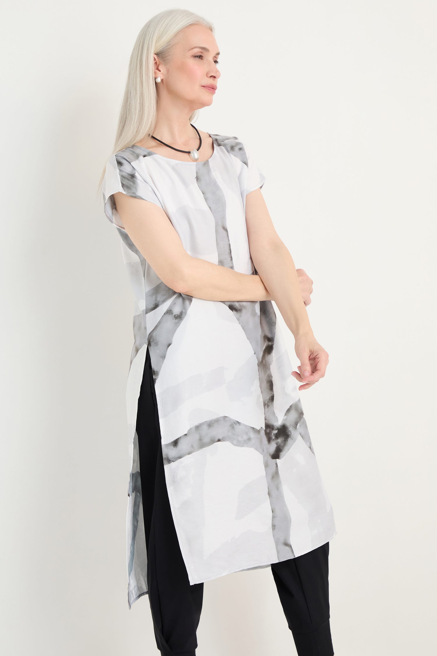 A woman with long gray hair models the Brushstroke Print Long Tunic featuring abstract black and gray patterns, paired with black pants and pearl earrings, standing against a plain light background.
