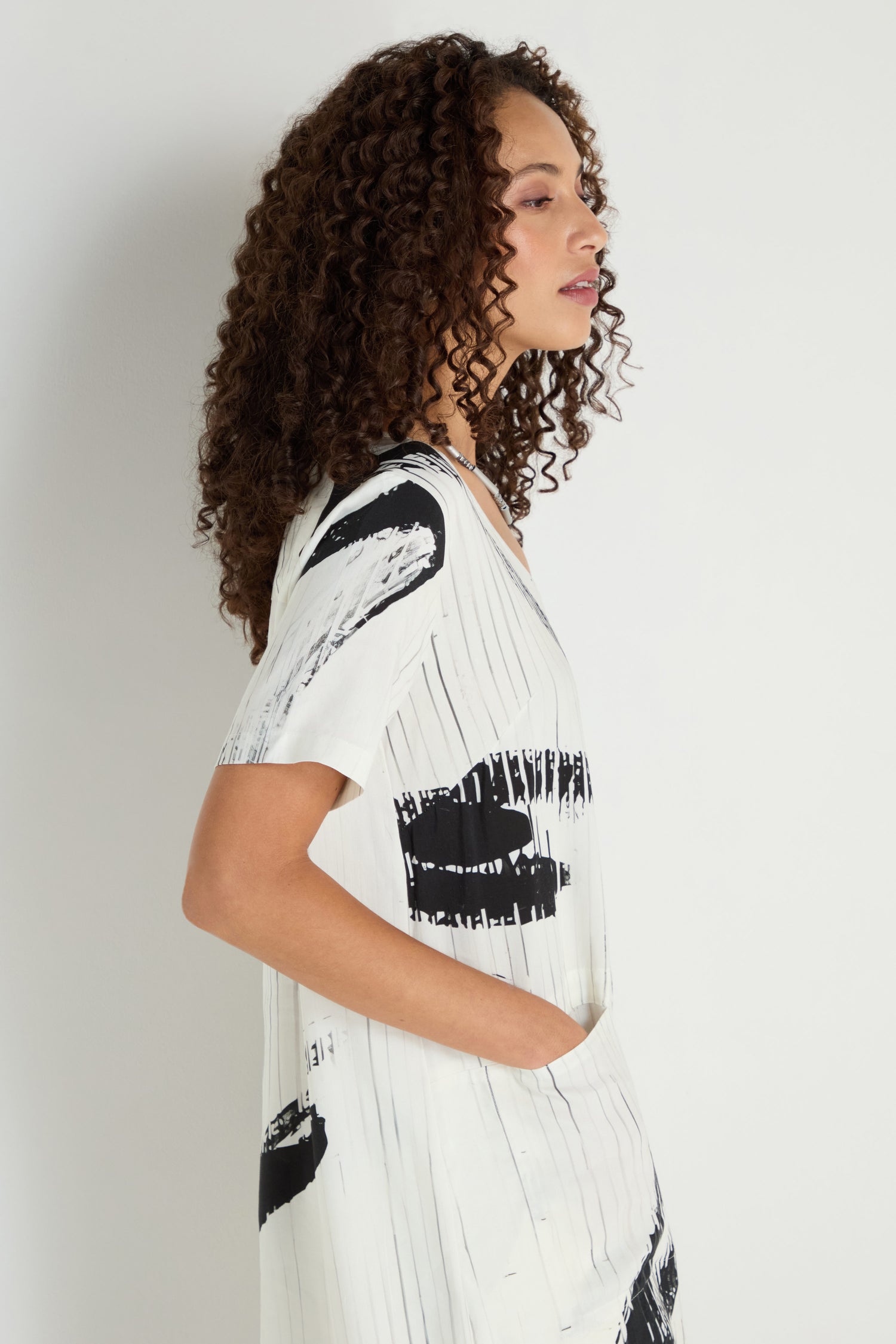 Woman with curly hair stands in profile, hand in pocket, wearing the Soft Stripe Midi Dress—a relaxed white midi dress with black abstract prints—against a plain light background.