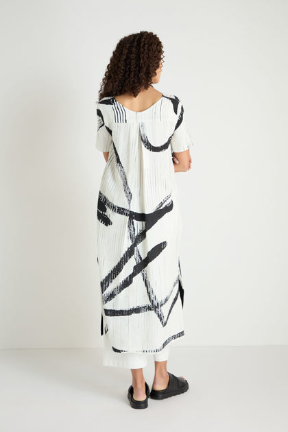 A person with curly hair, facing away, wears the Soft Stripe Midi Dress—a relaxed viscose linen blend with bold black abstract patterns—and black sandals, set against a plain white background.