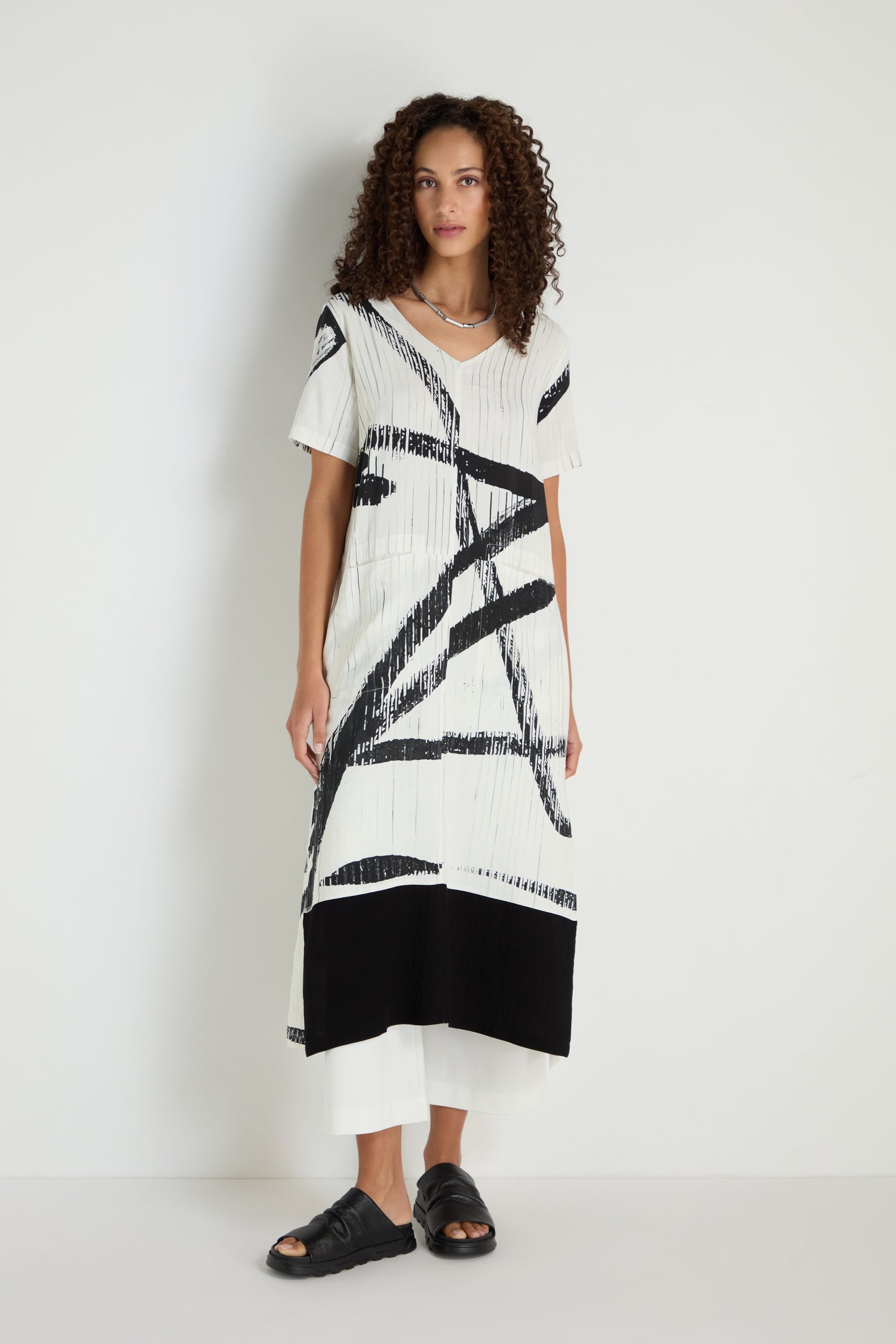 A woman stands against a white wall wearing the Soft Stripe Midi Dress, a relaxed white midi silhouette with black abstract patterns, styled with white pants underneath and black sandals.
