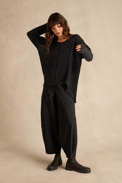 A woman stands against a plain beige background wearing the Black Seam Detail Scoop Neck Knit, paired with wide black pants and black boots, with one hand behind her head and looking downward.